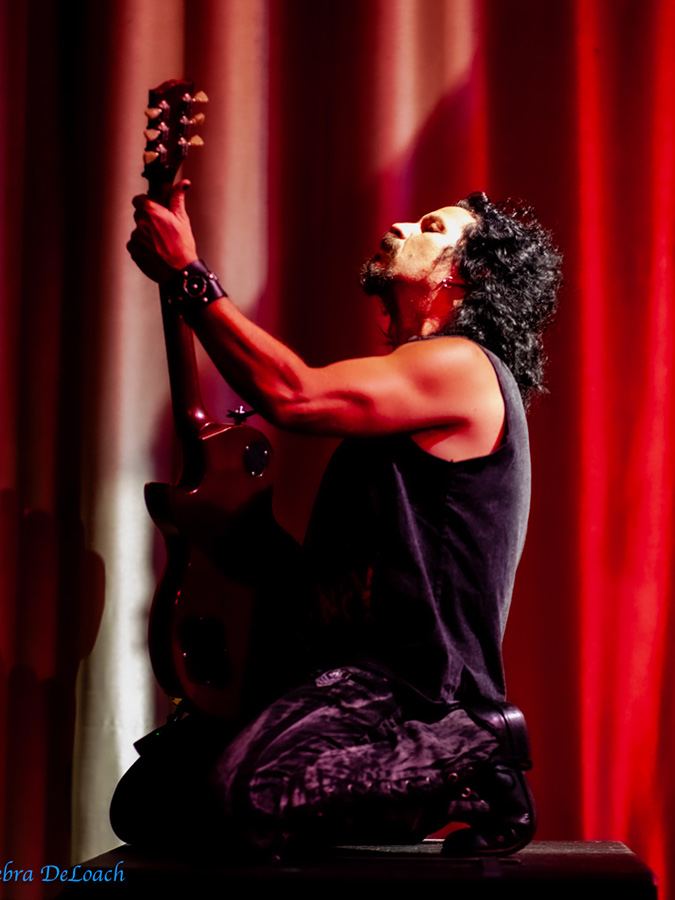 Mike Ruggirello playing a Gibson Les Paul kneeling in front of a red curtain